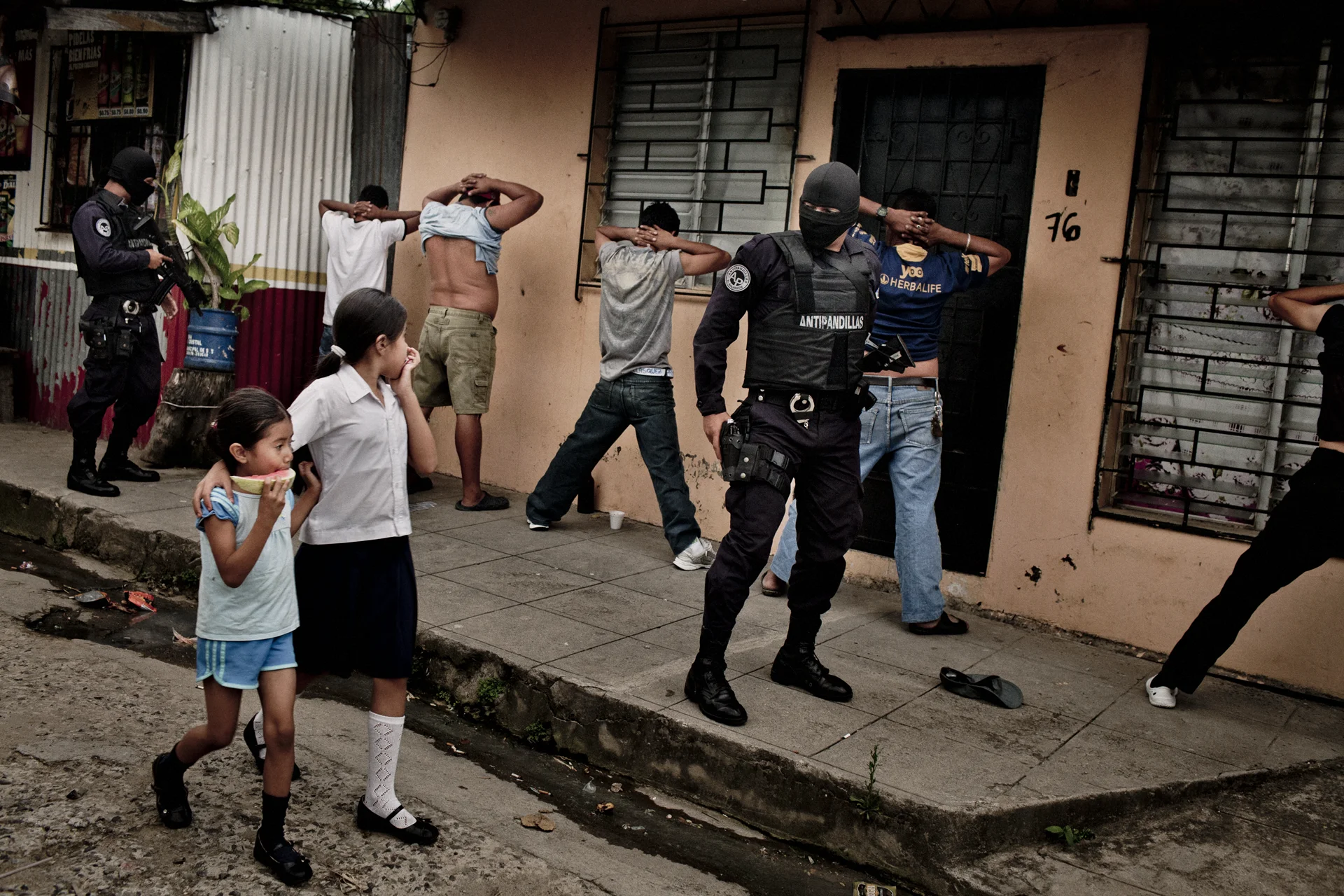 Policía antipandillas busca pandilleros mientras dos niñas caminan enfrente de ellos.
