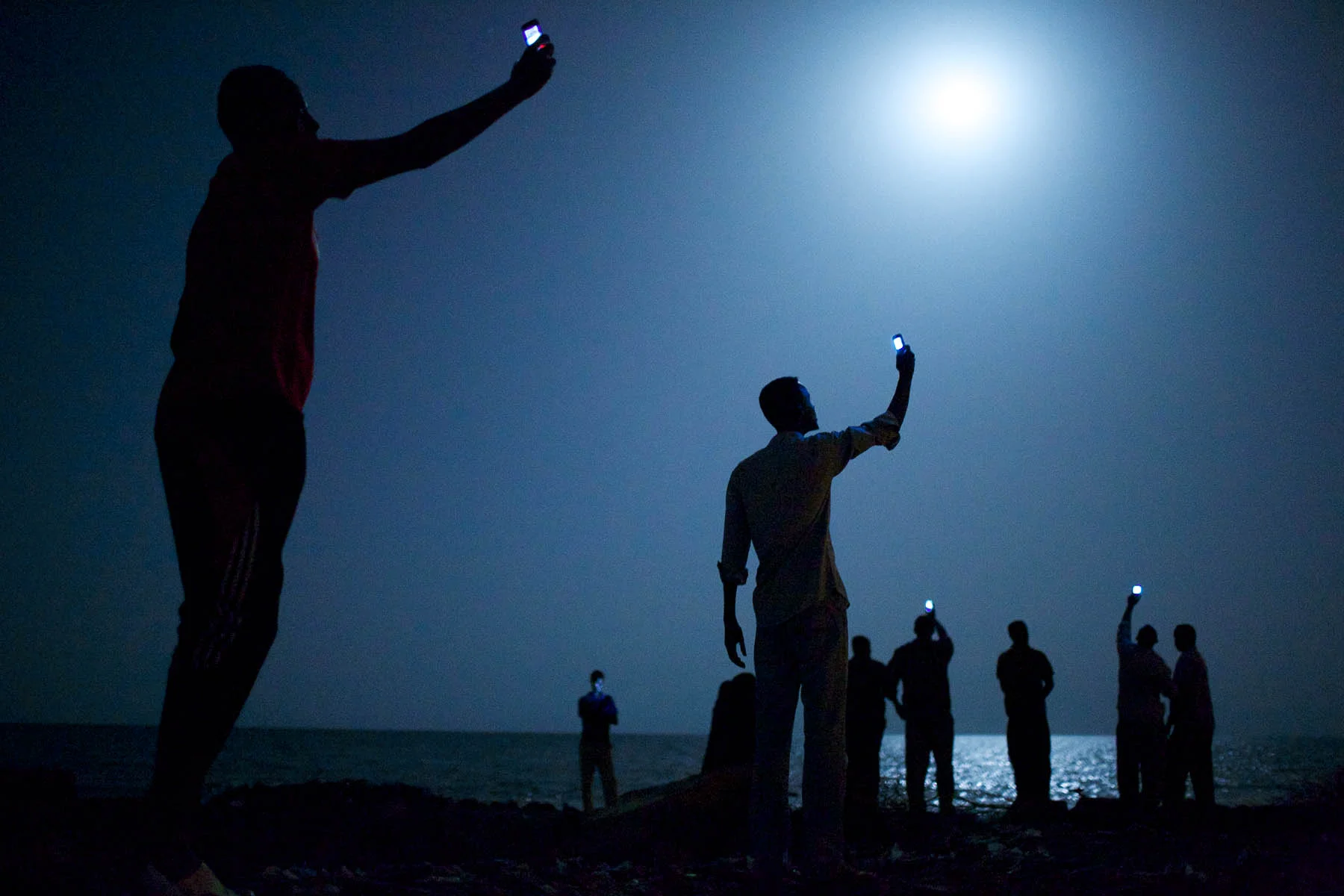 Migrantes africanos en la costa de la ciudad de Djibouti en la noche levantan sus teléfonos en un intento de captar una señal de la vecina Somalia, un vínculo con sus familiares en el extranjero.