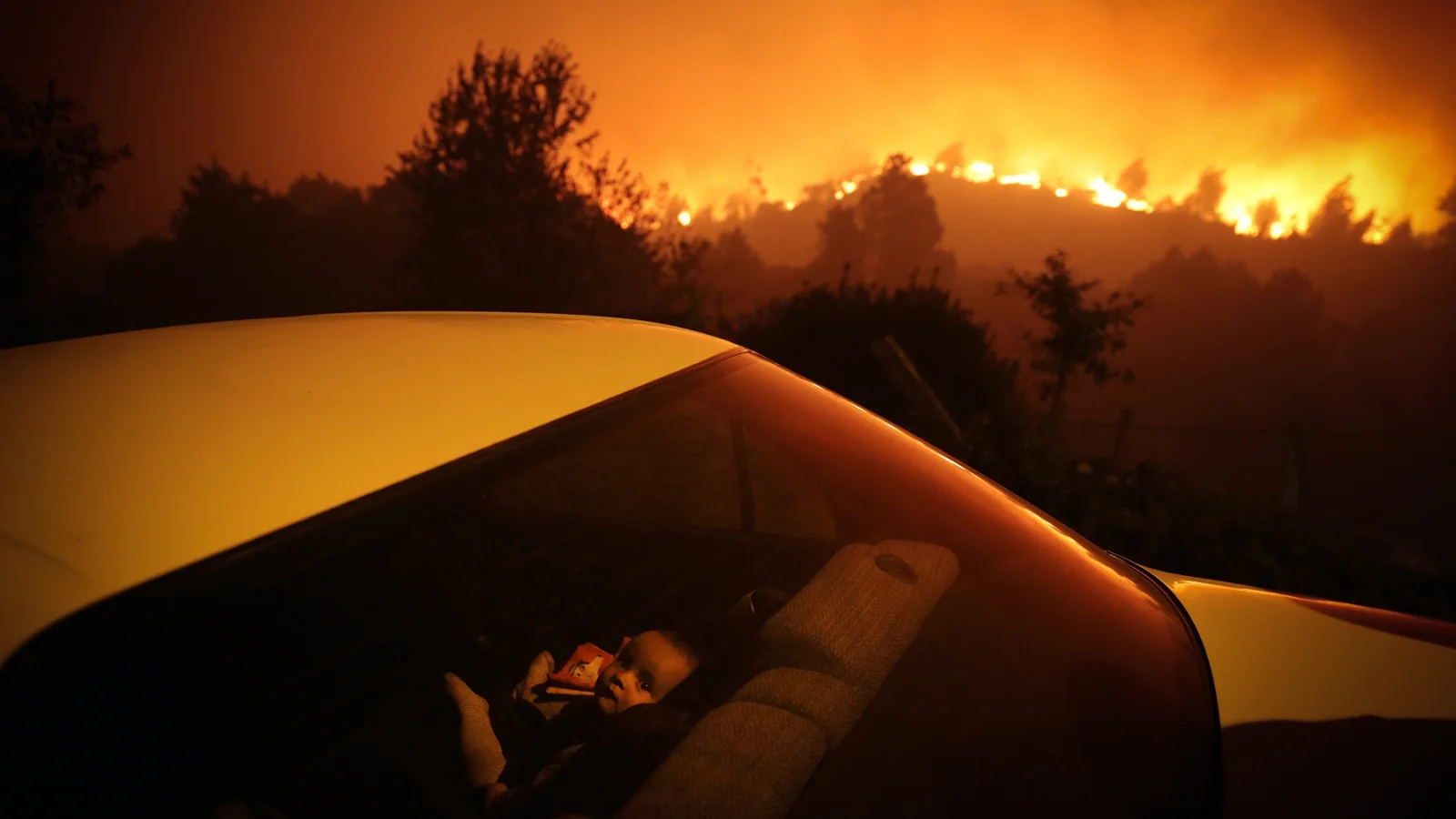 Um menino sentado dentro de um carro perto de um incêndio florestal.