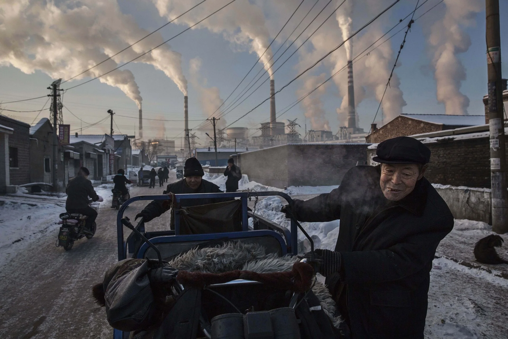 El humo sale de las chimeneas mientras los hombres empujan un triciclo por un vecindario al lado de una planta de energía a carbón en la provincia norteña de Shanxi.